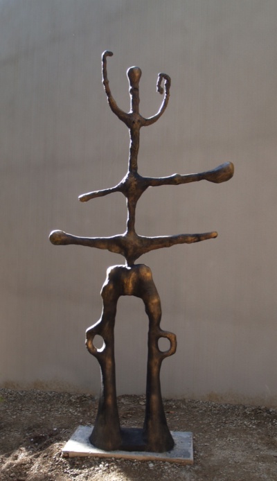 Escultura exempta de bronze: una figura totèmica de dues cames arquejades amb finestres rodones, tors de braços horitzontals i un cap petit amb tres tiges alçades. Fotografiada a l'exterior, contra una paret llisa.