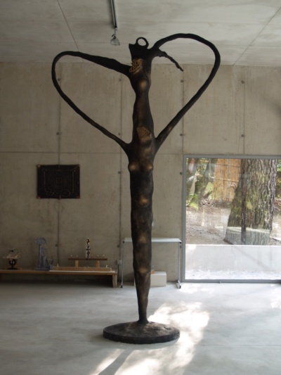 Escultura exempta: una figura femenina allargada de bronze fosc, amb els braços alçats i corbats formant un cor sobre el cap, damunt d'una base circular. Fotografiada al taller d'escultura, amb el bosc rere el vidre.